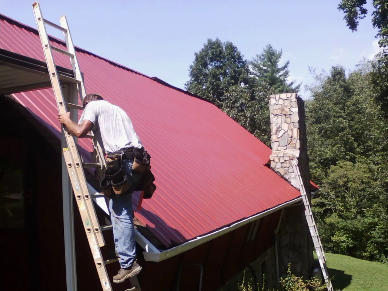 Roofer on a ladder beside a red metal roof and stone chimney — Soffit & Fascia Repair by Roofing Brothers in Archbold